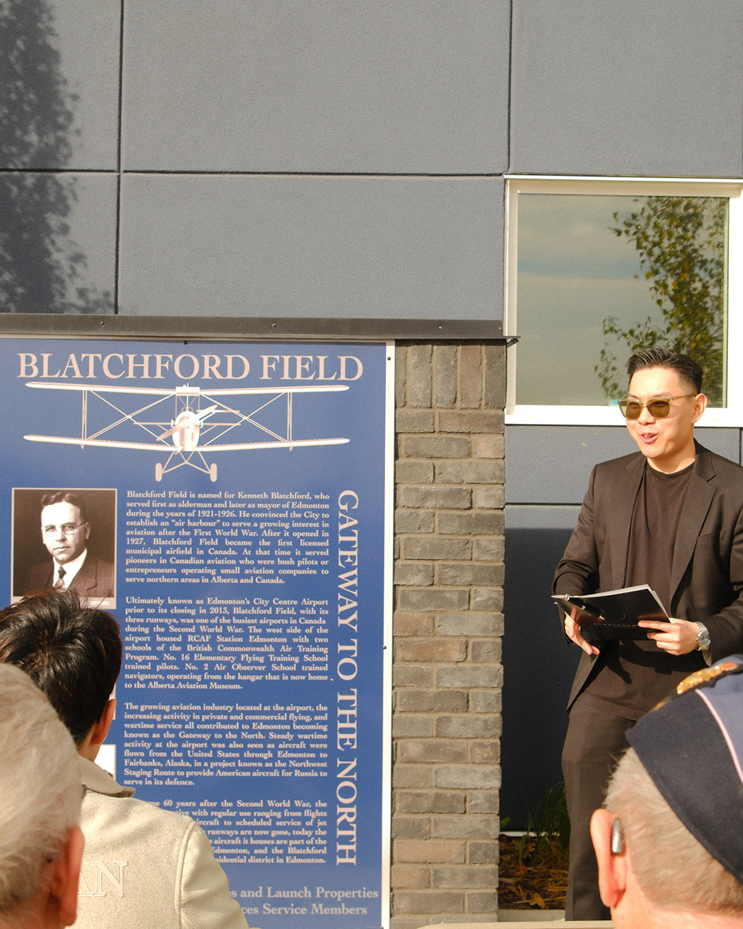 A man stands next to a plaque while speaking to a seated audience.